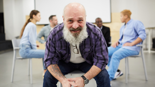 Man sitting on chair leaning forward while a 12-step group session happens behind him